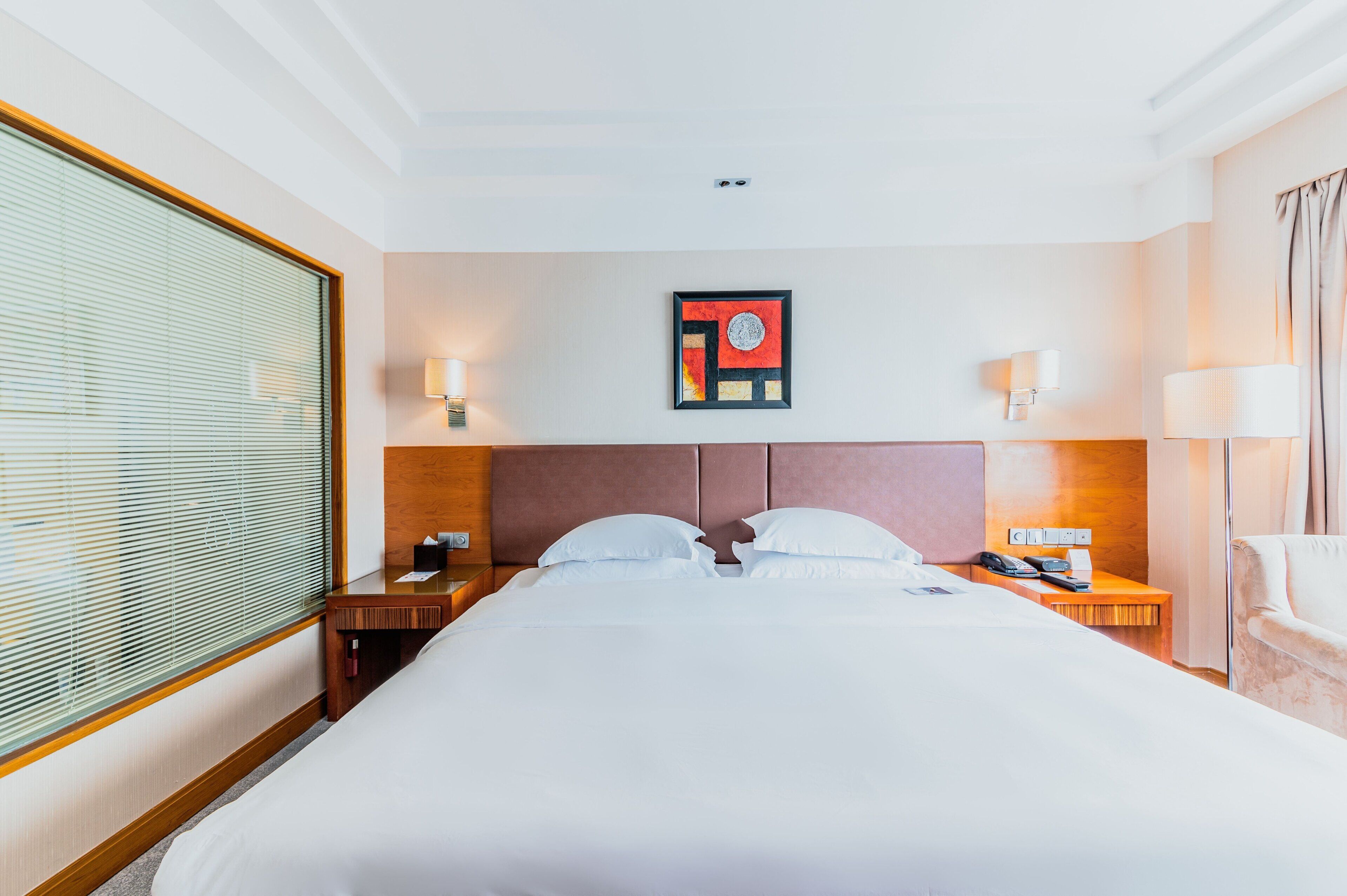 Una habitación de hotel con una cama grande, mesitas de noche, una obra de arte colgada en la pared y una ventana con persianas.