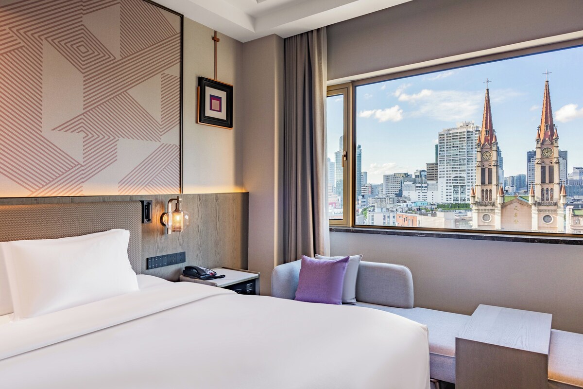 Ein Hotelzimmer mit einem großen Bett, einem Schreibtisch und Blick auf eine Stadtlandschaft, darunter ein Kirchturm.