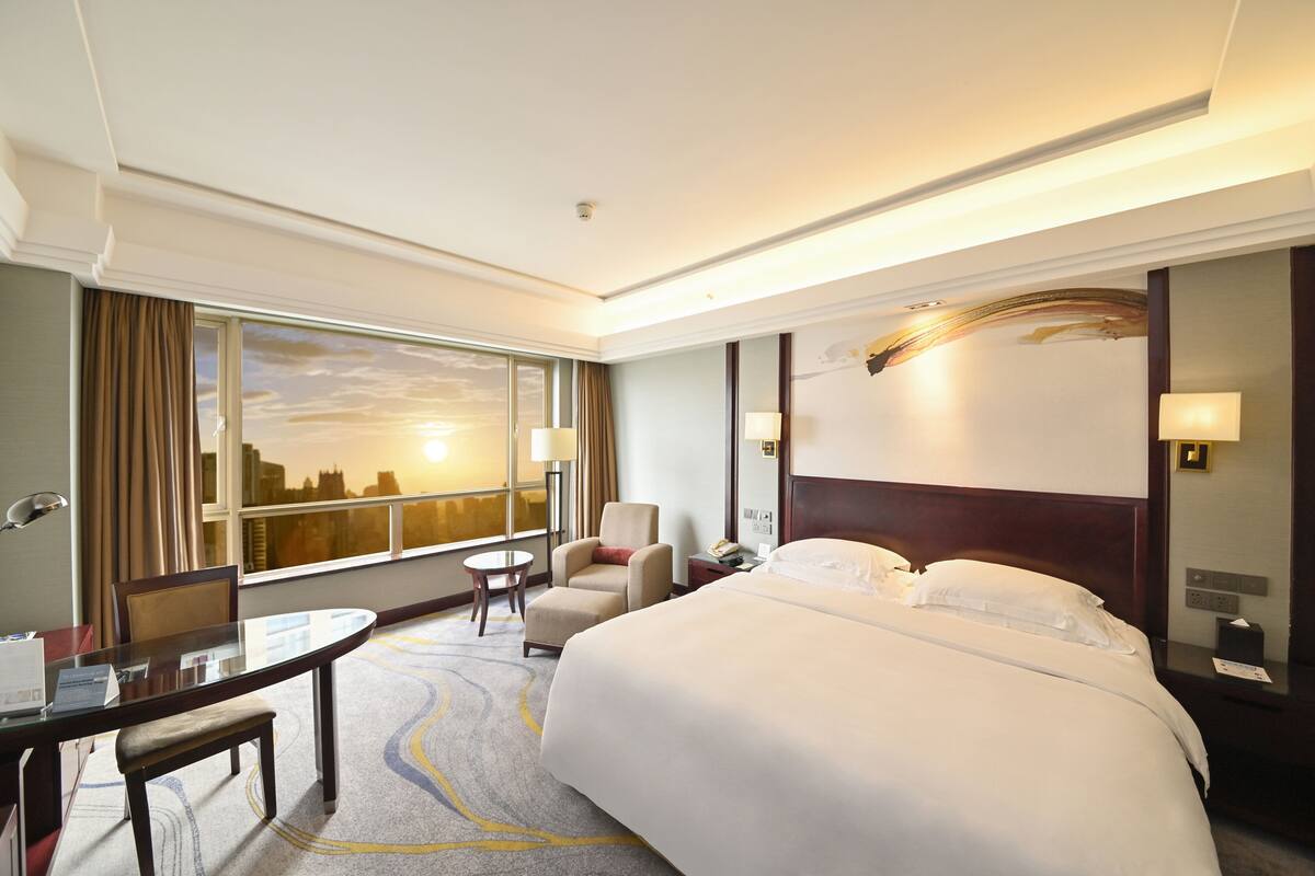 Habitación de hotel con una cama grande, un escritorio, una silla, una mesita y vistas de la ciudad a través de la ventana.