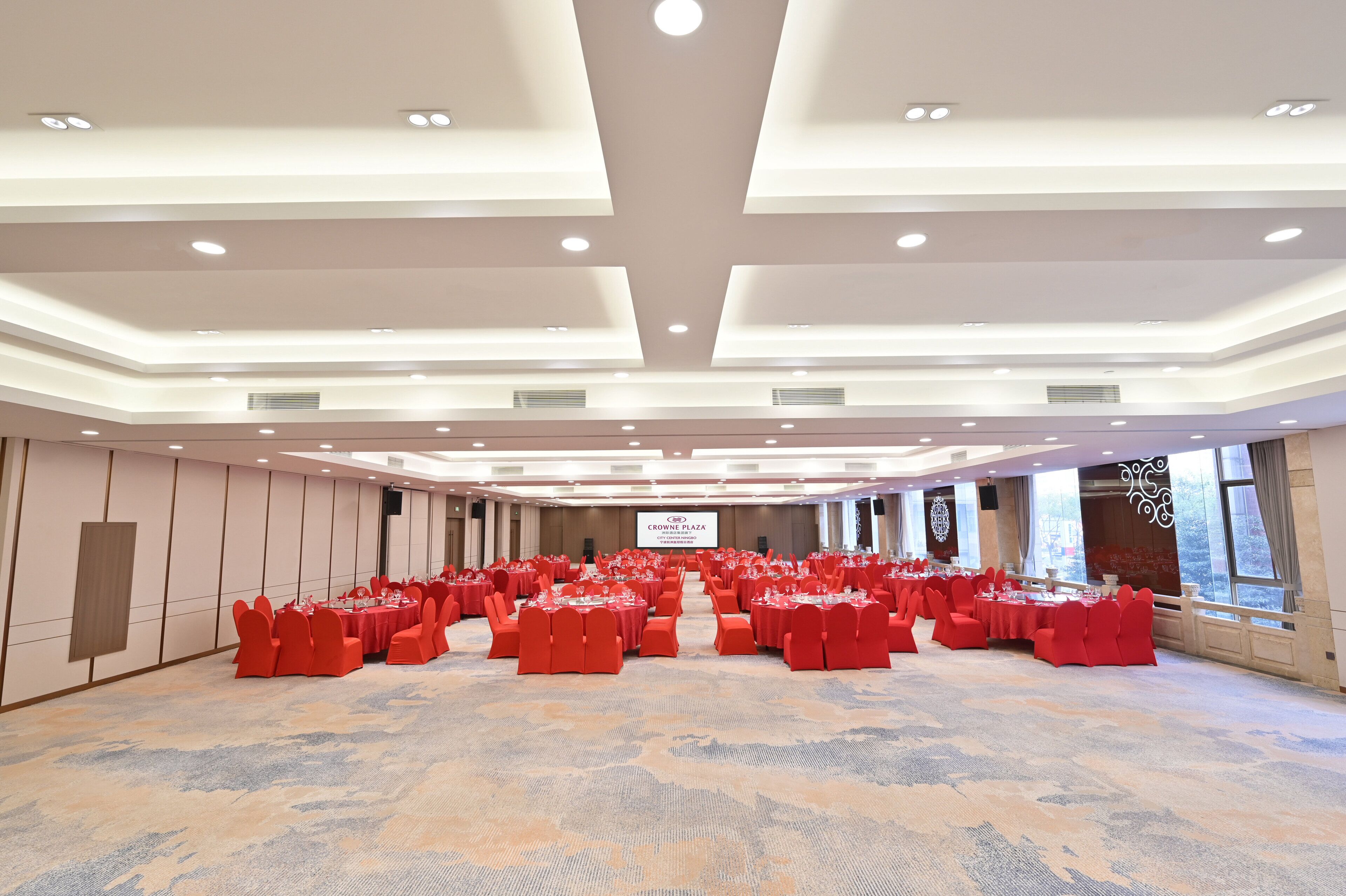 Un amplio salón de conferencias con filas de sillas y mesas rojas, un atril y una pantalla grande.