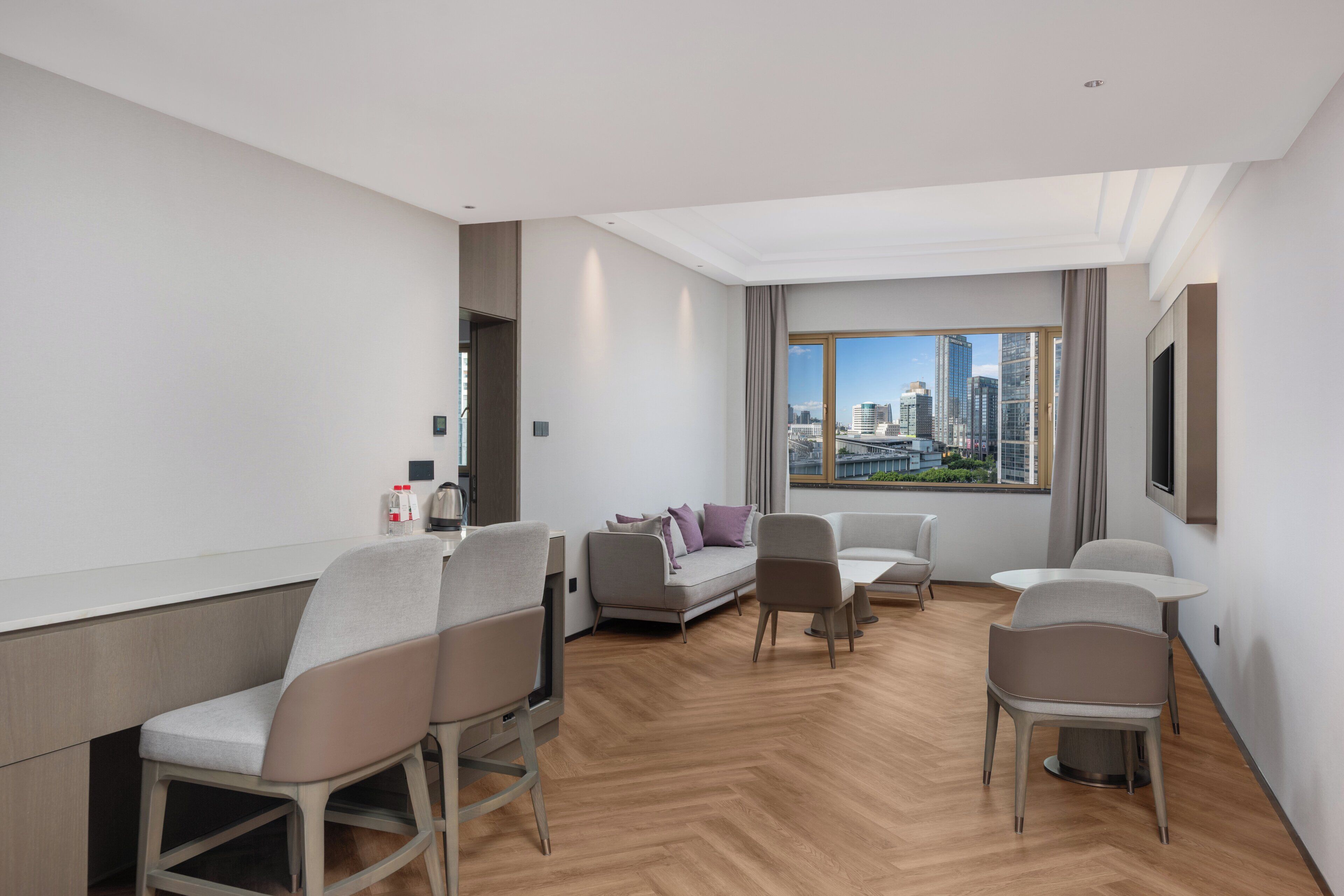 A modern hotel room with a city view, a sofa, chairs, and a dining table.