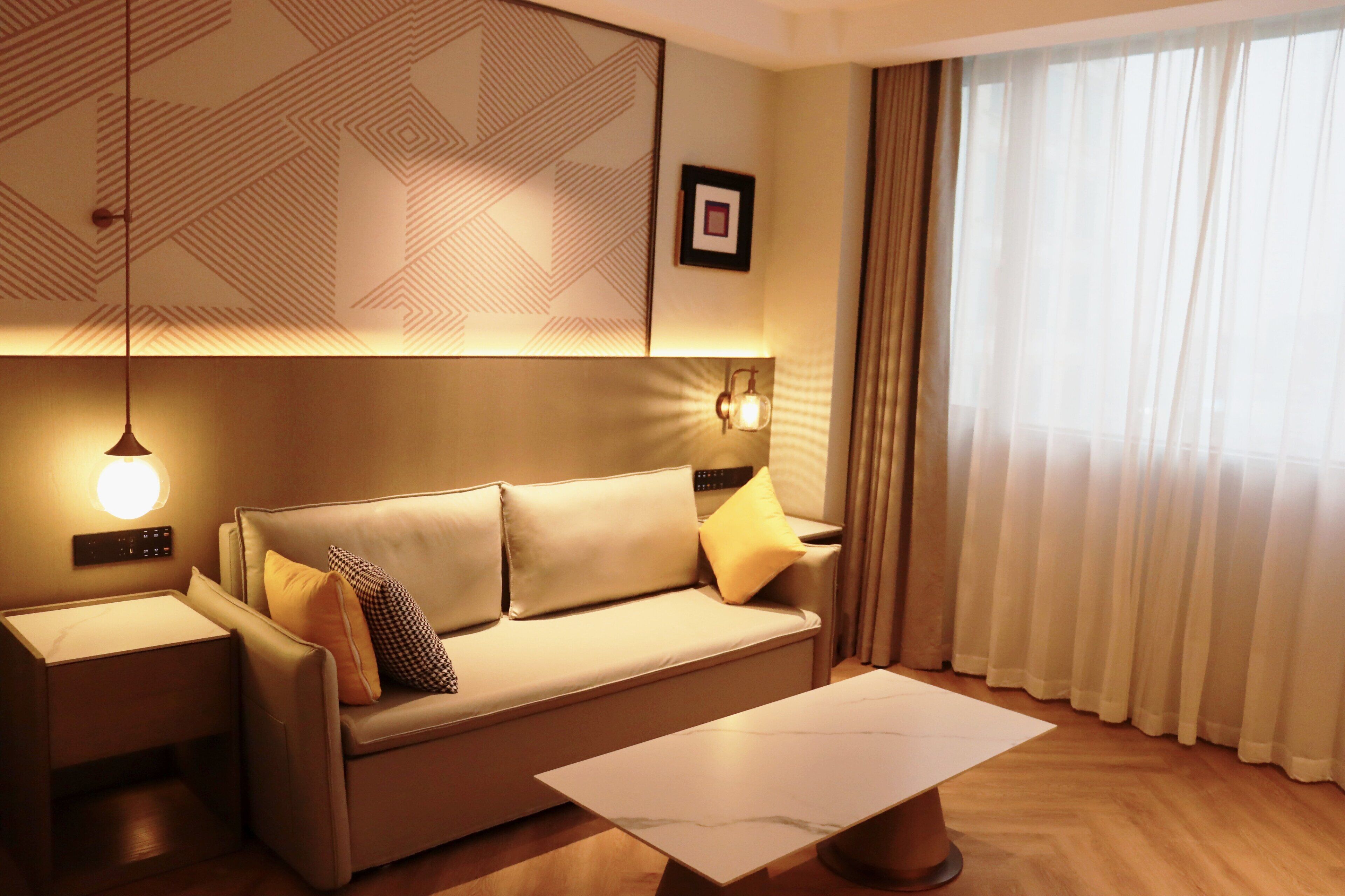Ein modernes Hotelzimmer mit einer Couch, einem Couchtisch, einer Wandleuchte und einem großen Fenster mit Vorhängen.