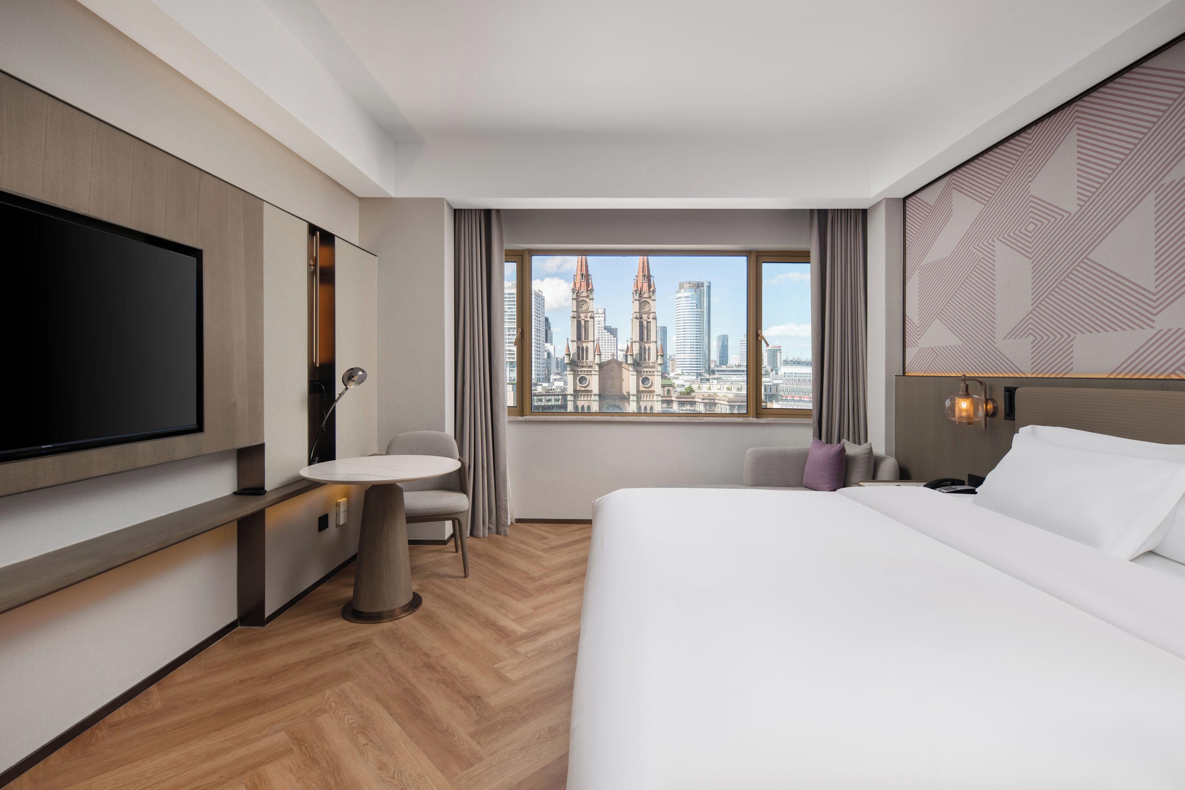Habitació d'hotel moderna amb un llit gran, una televisió de pantalla plana, una taula rodona petita, una cadira i vista a un paisatge urbà amb edificis alts.