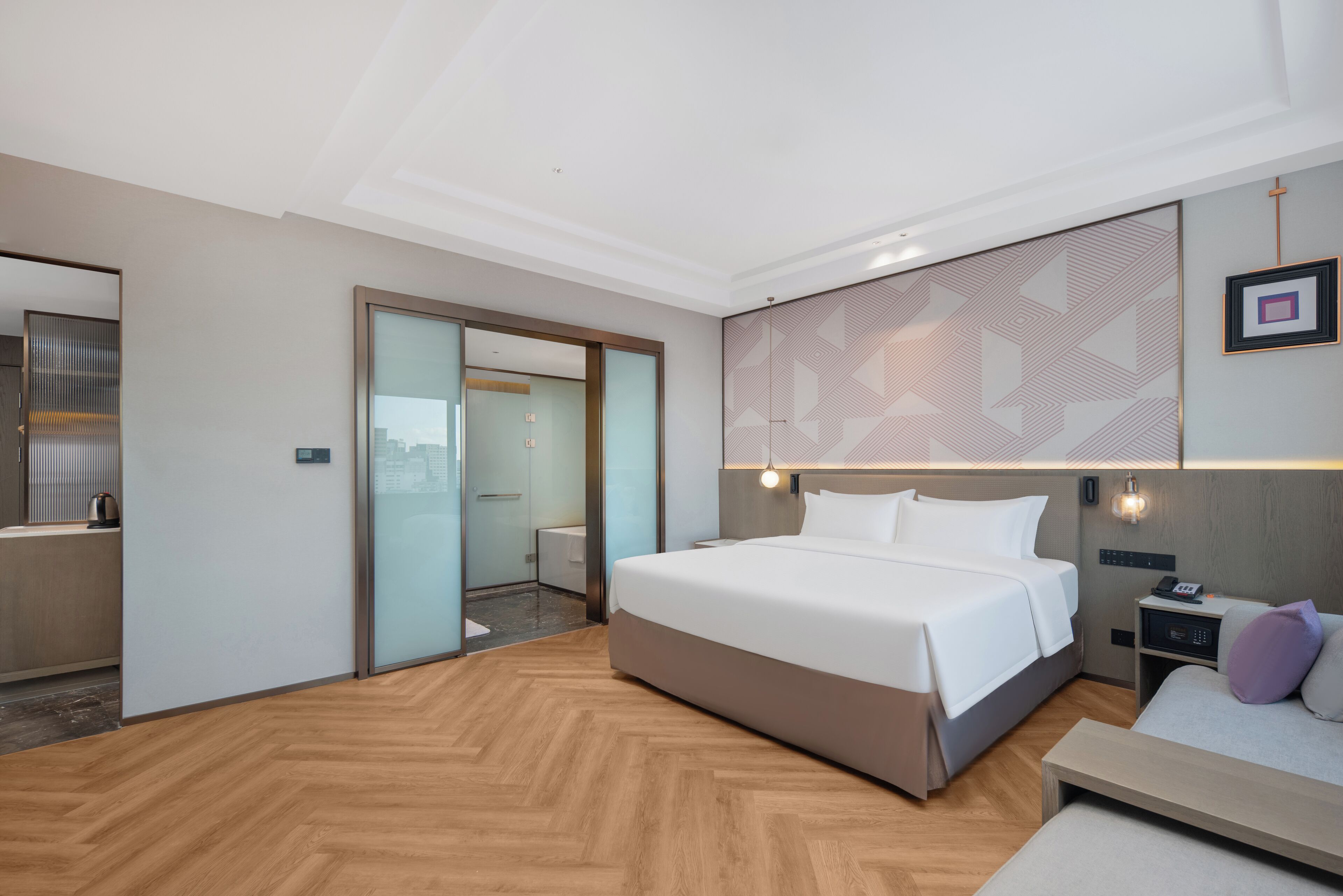 A modern hotel room with a large bed, a bedside table, a nightstand, and a view of the city through a glass door.