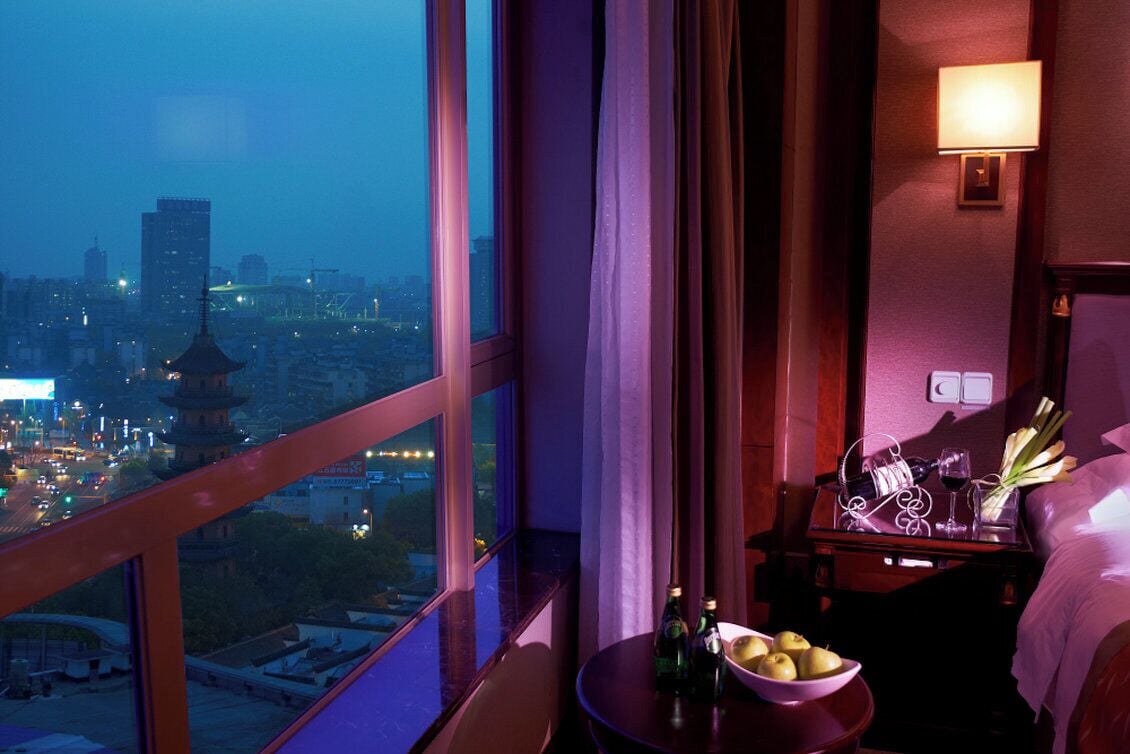Habitación de hotel con un ventanal grande que ofrece vistas a la ciudad, una mesita de noche con un frutero y botellas, y una lámpara de noche.