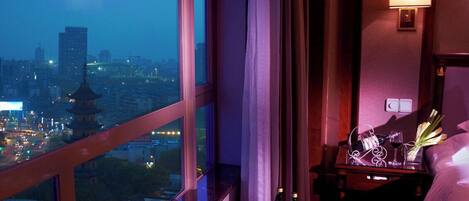 Habitación de hotel con un ventanal grande que ofrece vistas a la ciudad, una mesita de noche con un frutero y botellas, y una lámpara de noche.