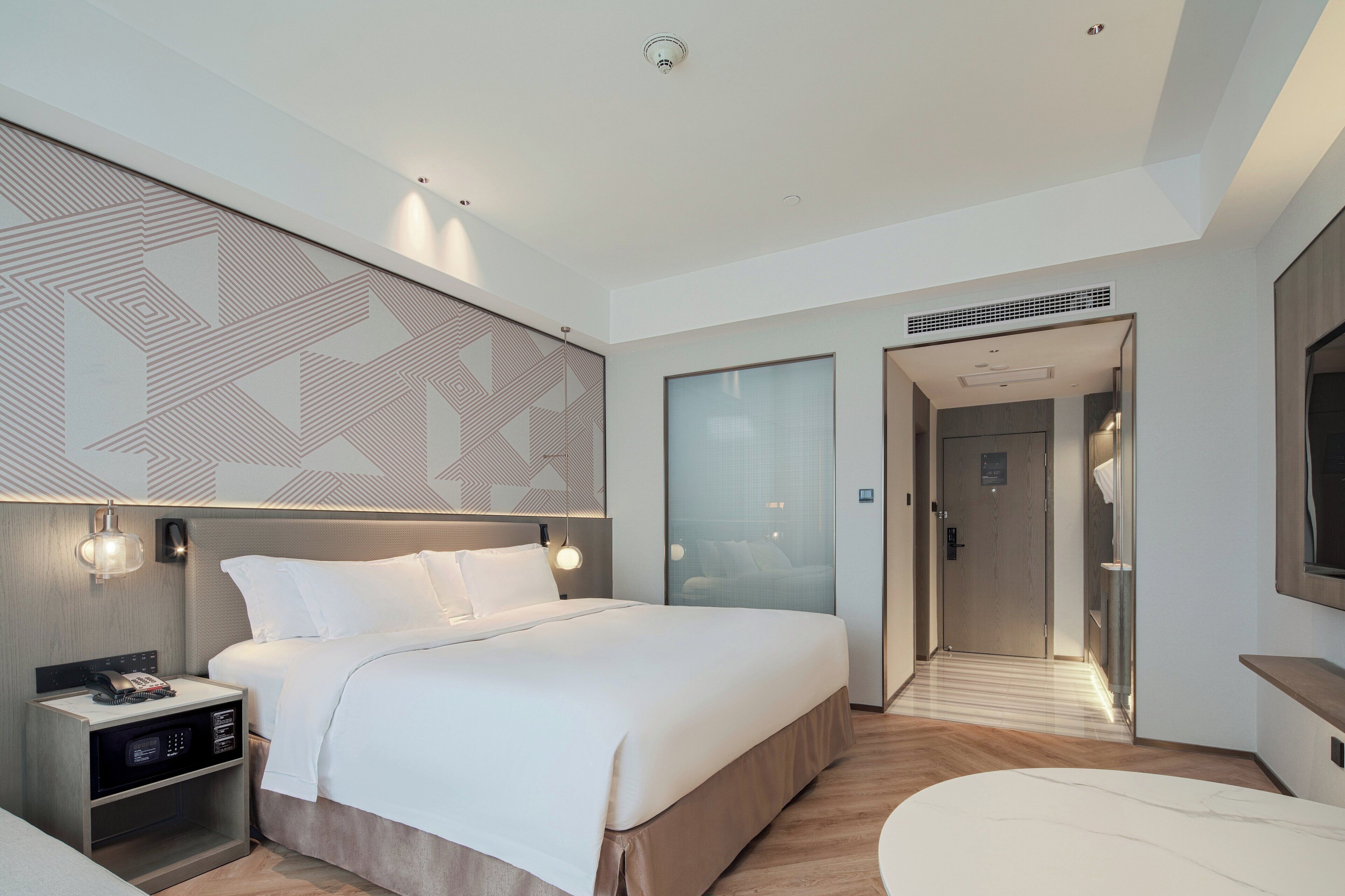 A modern hotel room with a large bed, bedside table with a phone and charger, a wall-mounted light, and a large mirror.