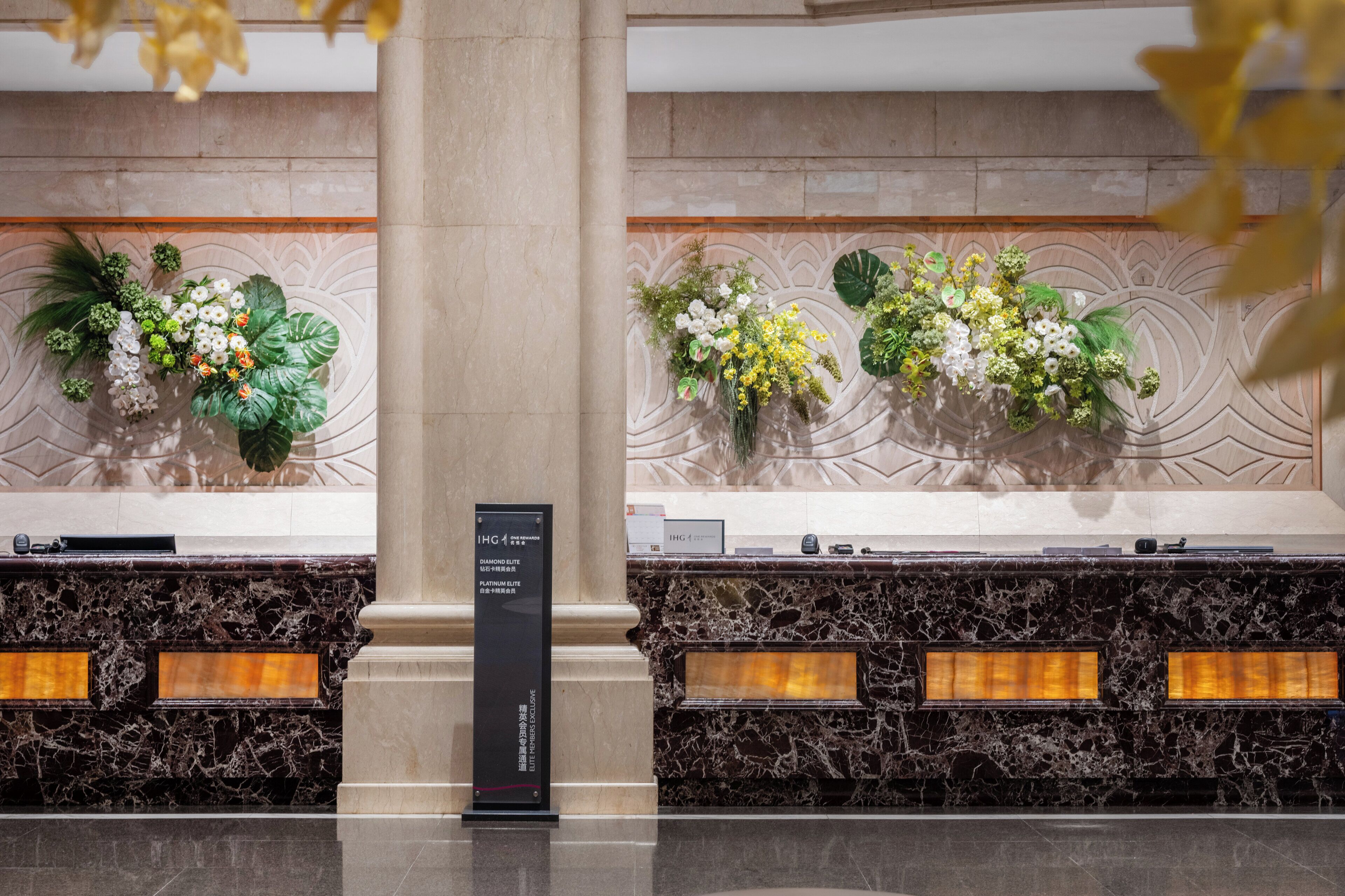 Una recepción con mostrador de mármol, dos arreglos florales y un letrero que indica el nombre del hotel y sus servicios.
