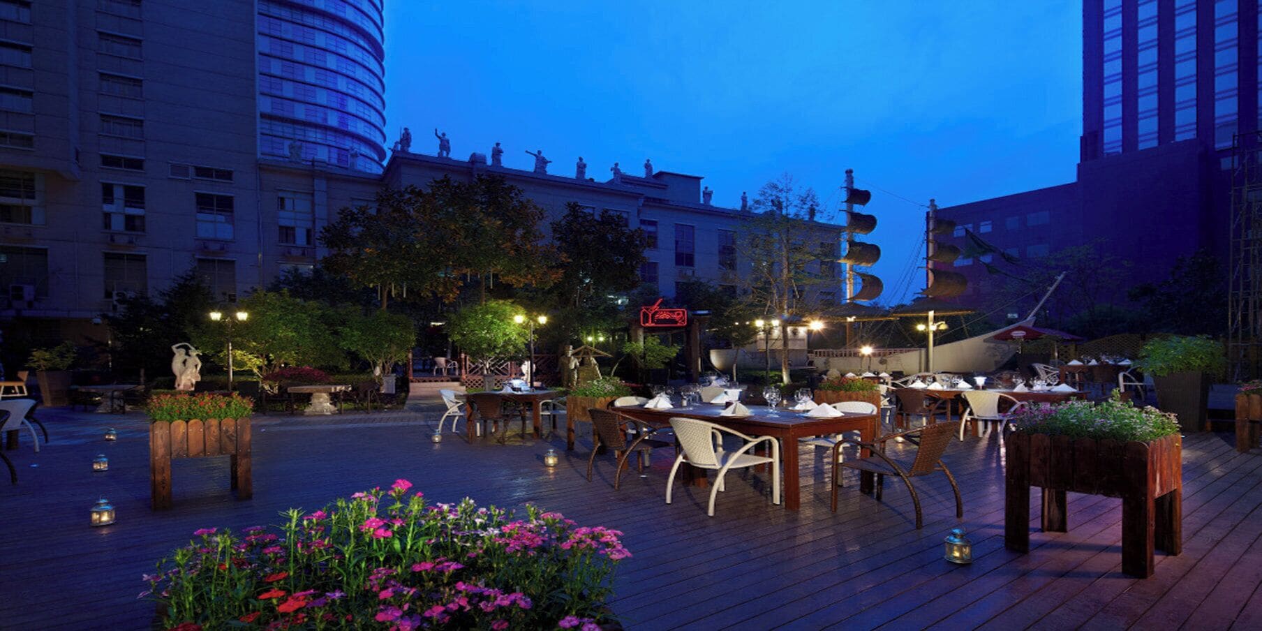 Un área de comedor al aire libre con mesas y sillas, rodeada de follaje y detalles decorativos.