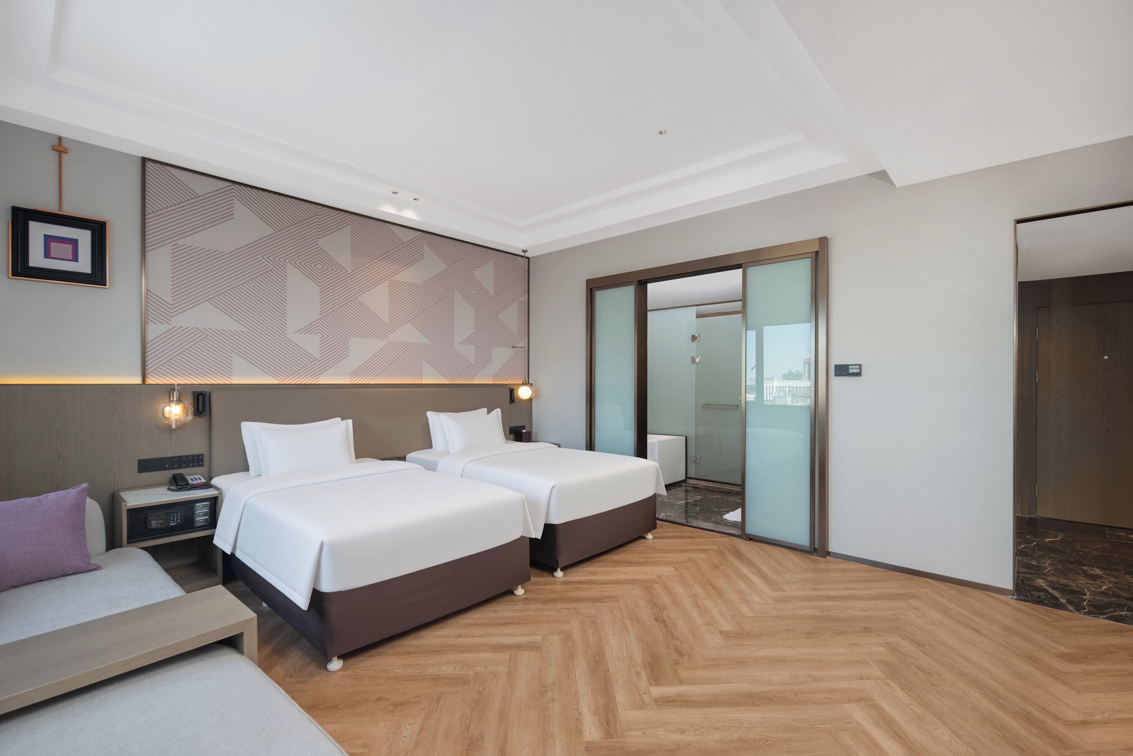 A hotel room with two beds, a sofa, a bedside table with a phone, a wall-mounted light, and a patterned wall.