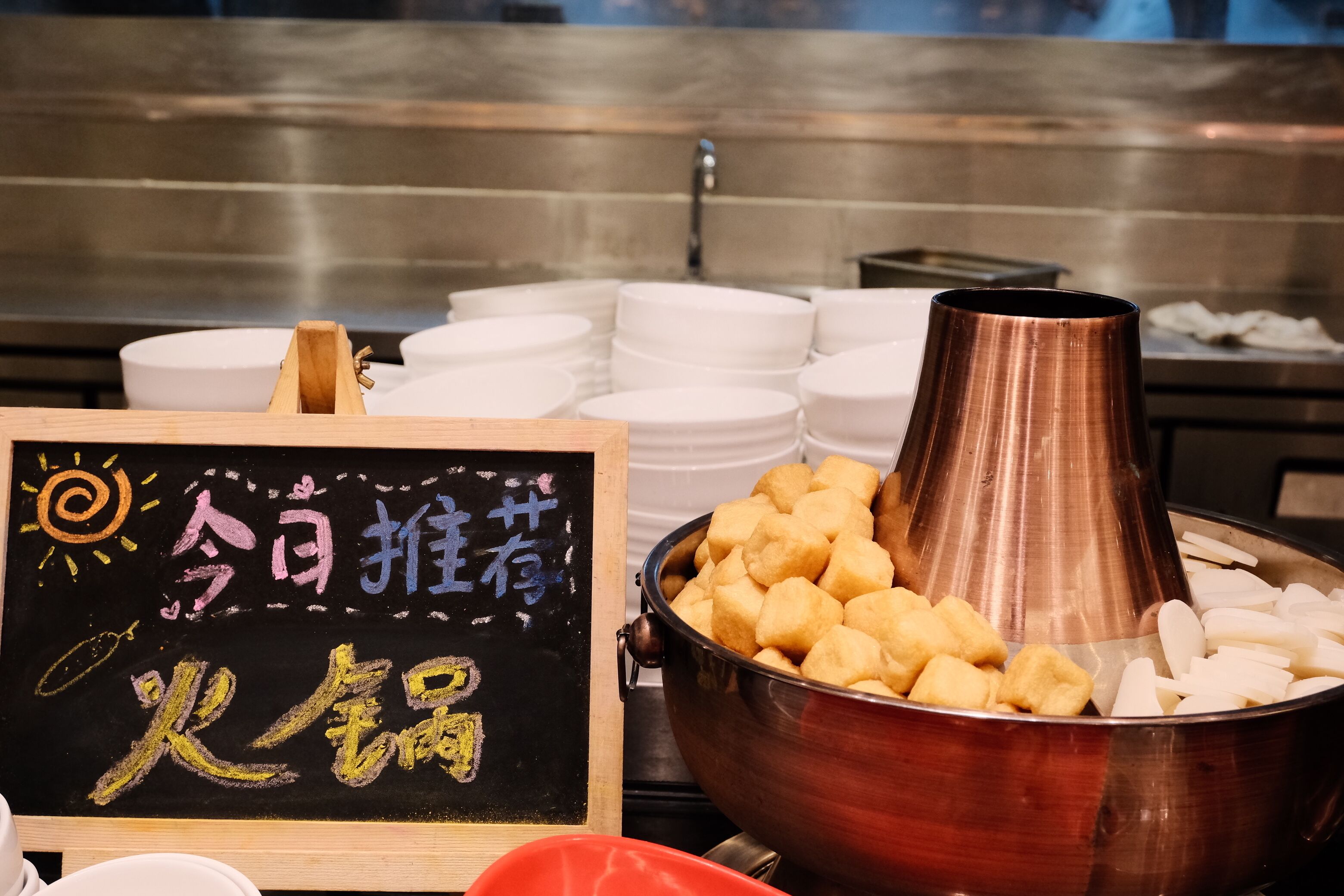 A buffet with a sign in a foreign language, a large copper pot, and stacked white plates.