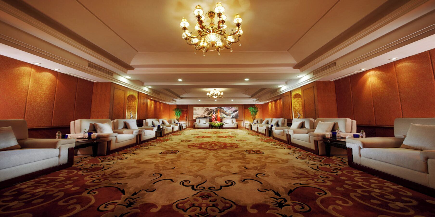 A hotel lobby with a long, ornate carpet, rows of seating, and a chandelier.