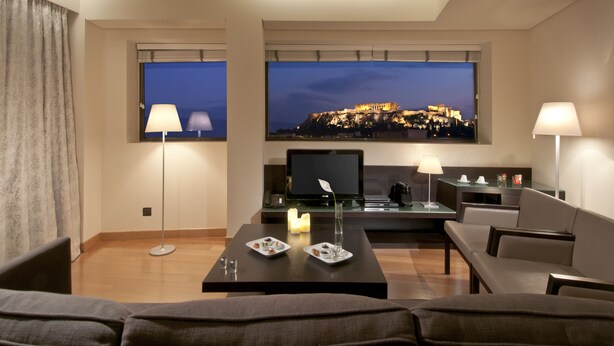 Una habitación de hotel moderna con un televisor de pantalla plana grande mostrando un paisaje urbano, un área de descanso acogedora con una mesa de centro, y vistas a un edificio histórico.