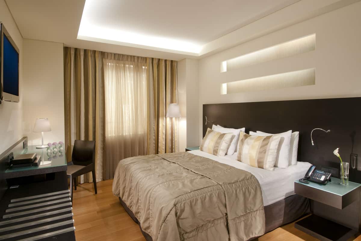 A modern hotel room with a large bed, a desk with a lamp, a chair, and a bedside table with a phone and vase.