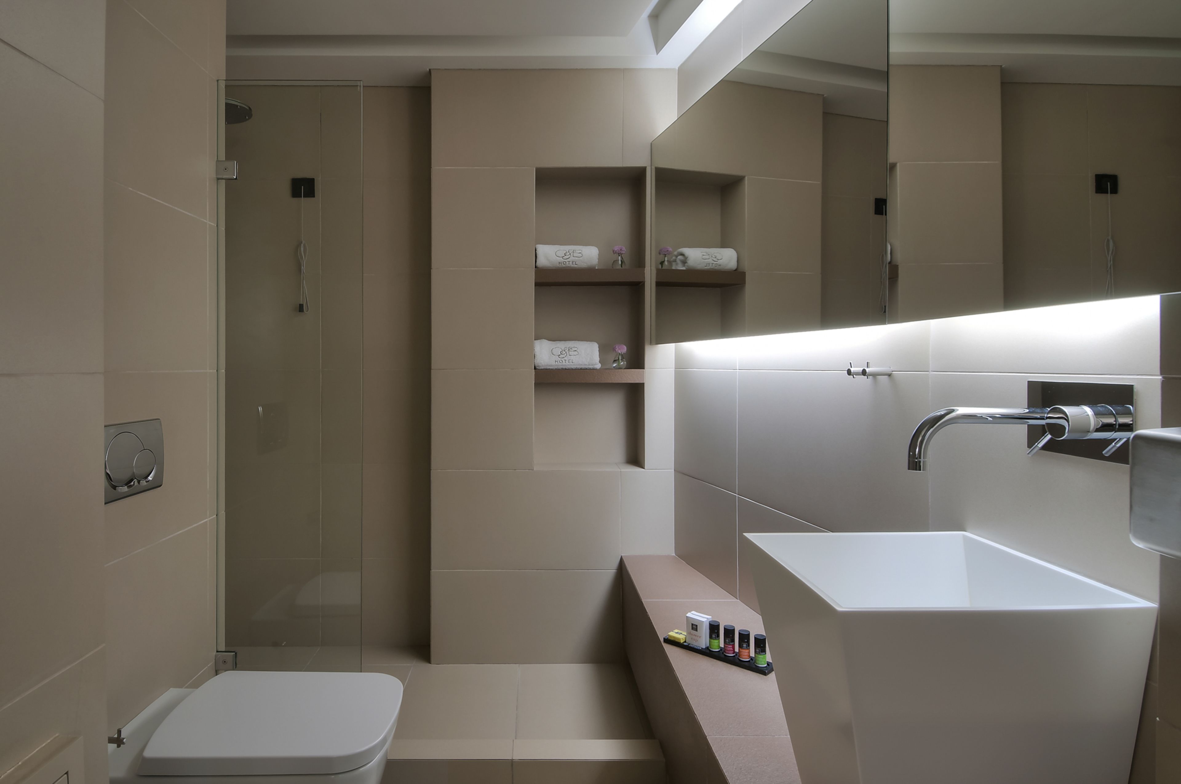 A modern bathroom with a glass-enclosed shower, a white sink, and a toilet. Shelves with towels and toiletries are visible.