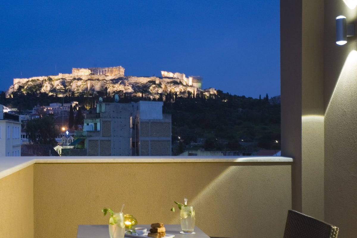 A balcony with a table and chairs, overlooking a historic cityscape with ancient ruins at night.