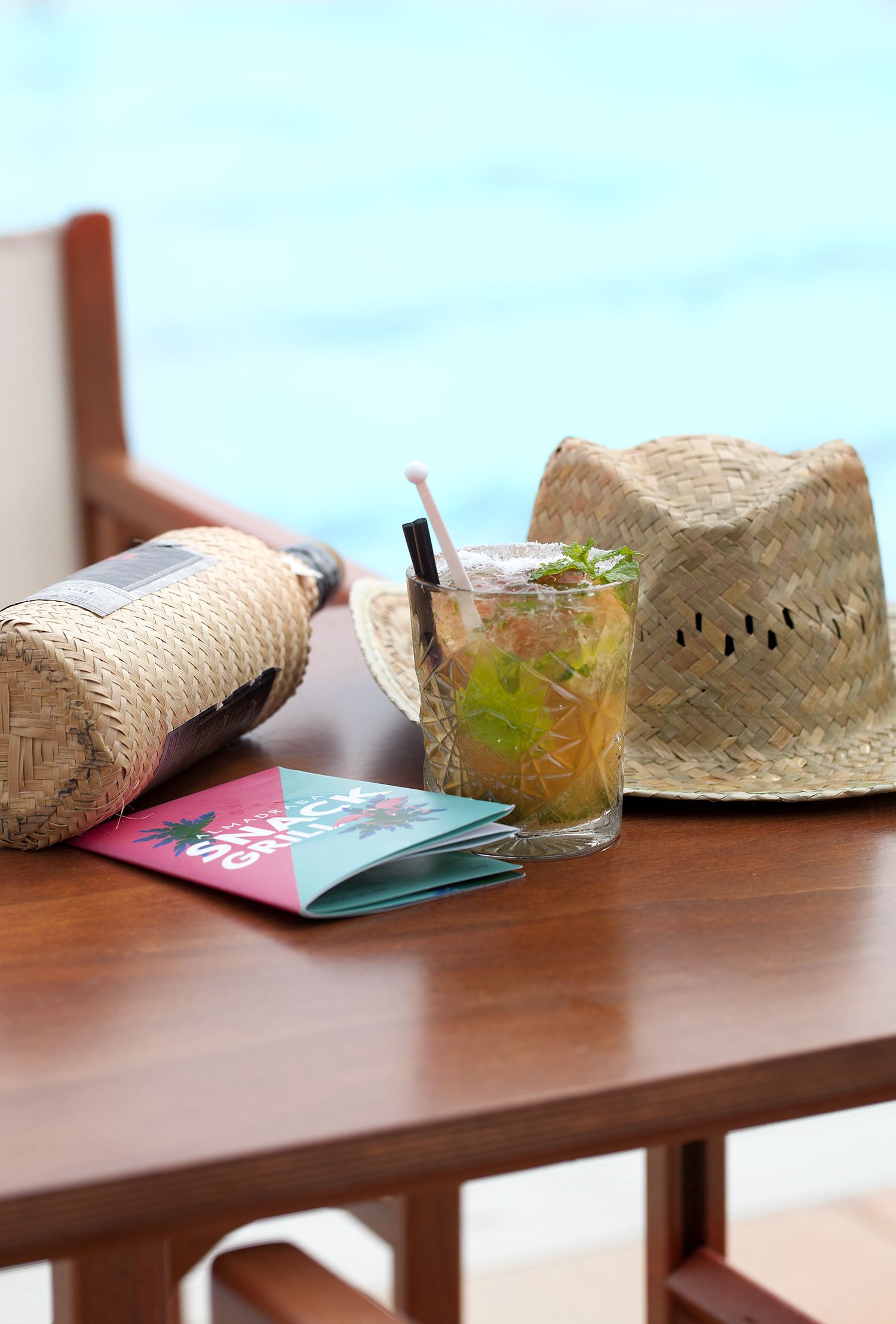 A wooden table with a straw hat, a bottle, and a cocktail glass with a straw.