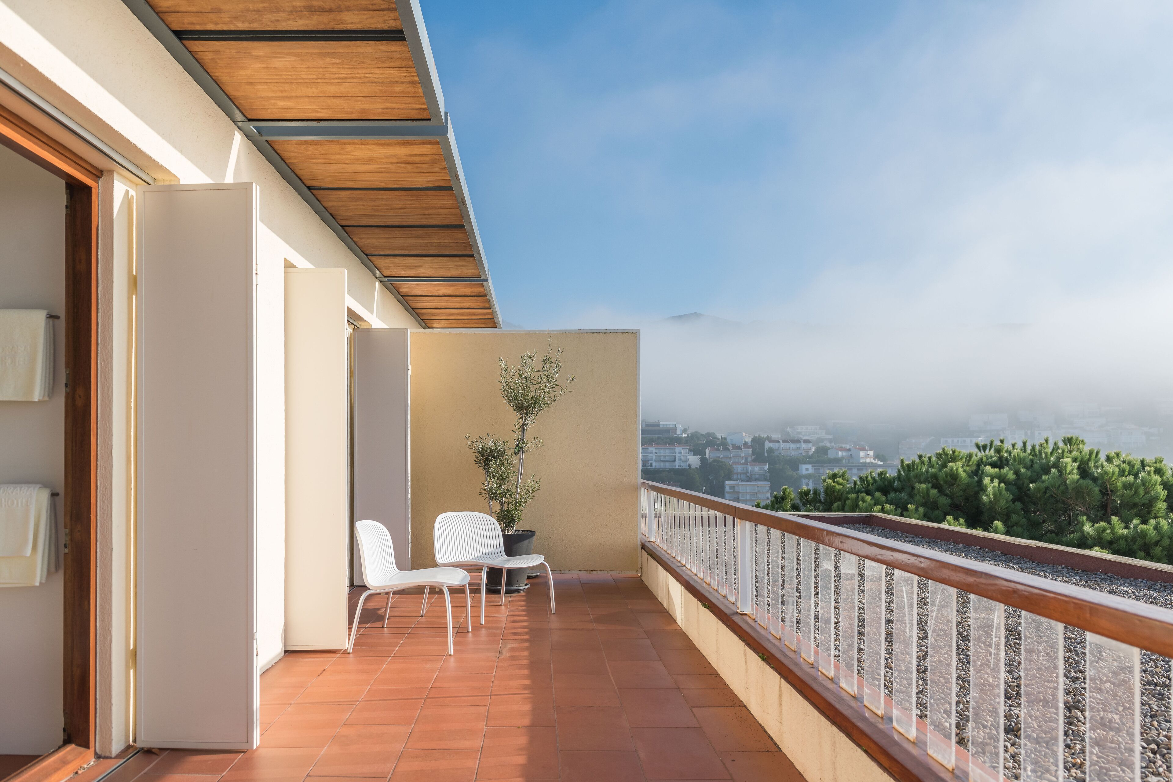A balcony with two white chairs and a potted plant, overlooking a cityscape with trees and a cloudy sky.