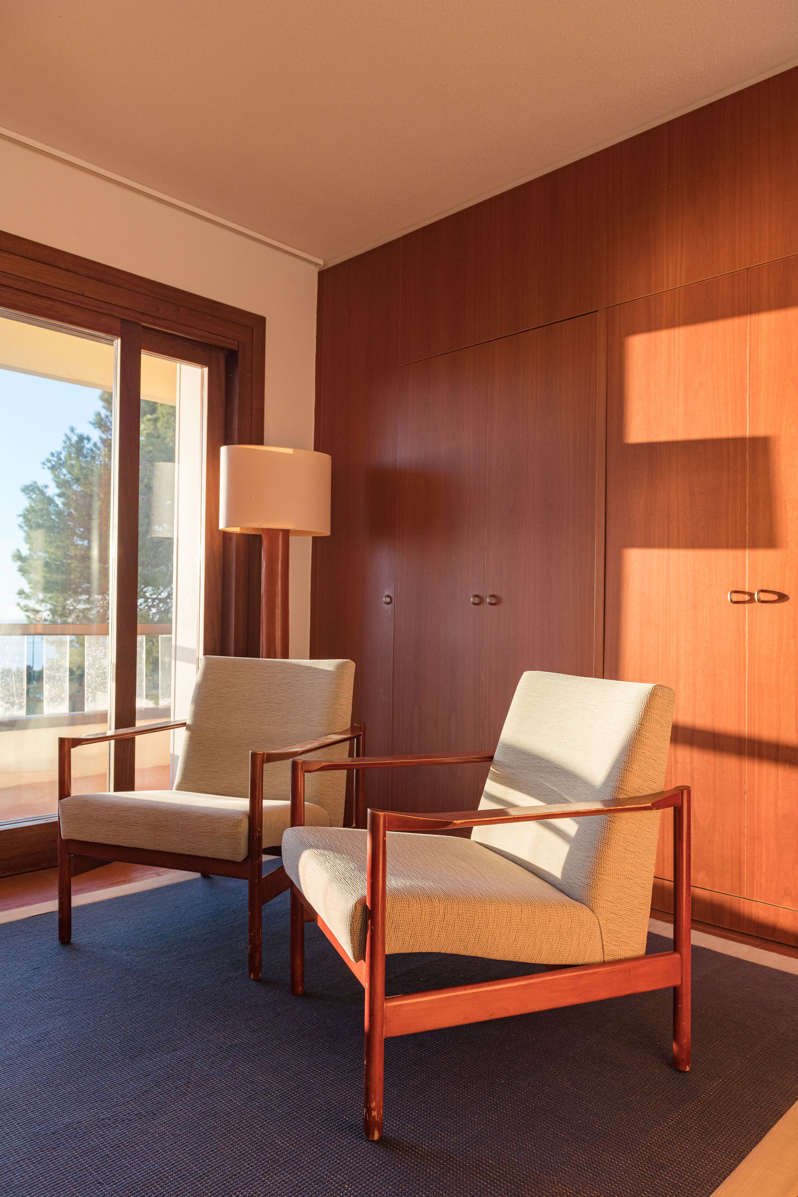 A room with two wooden armchairs, a large window, and a wall-mounted lamp.