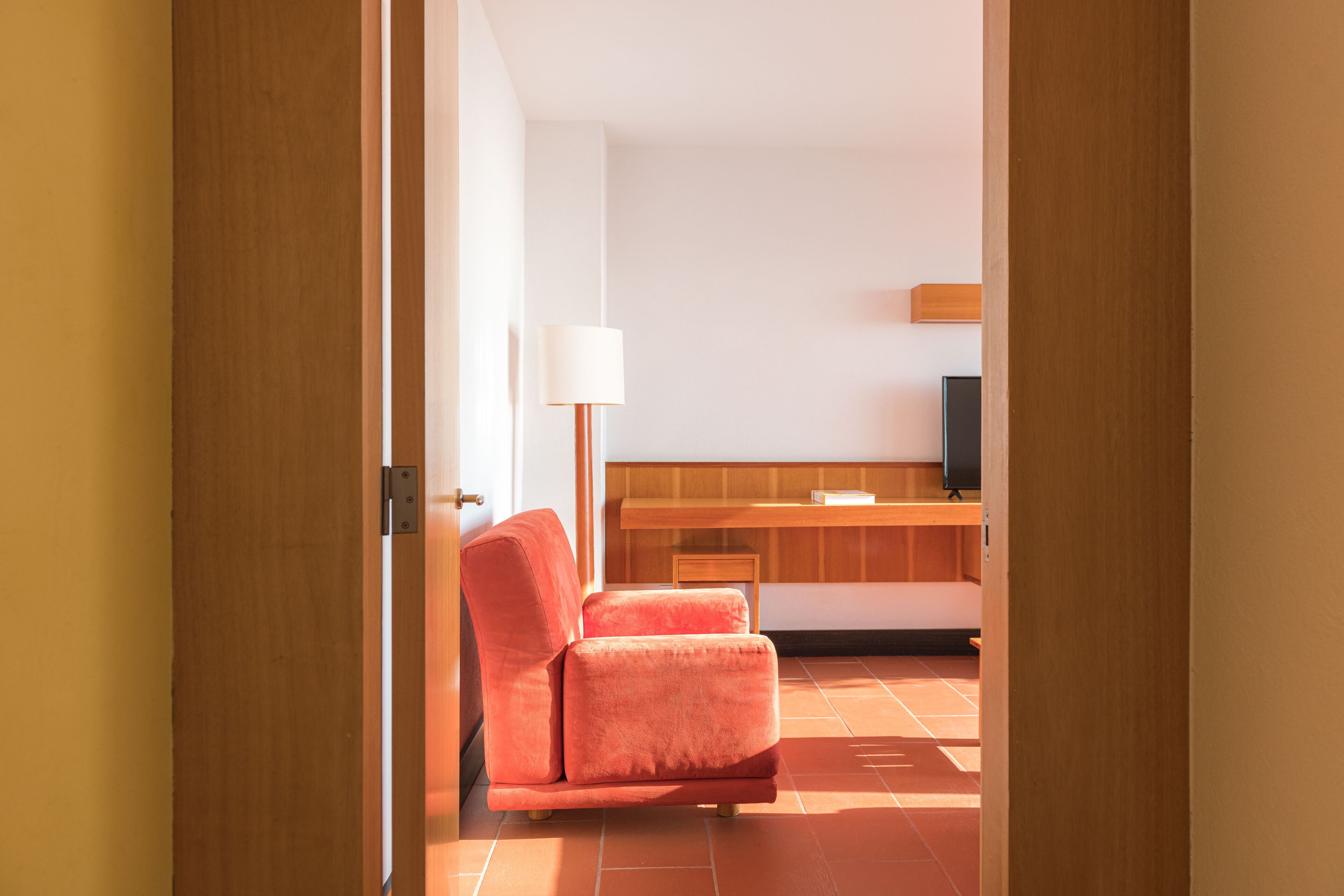 A modern room with a wooden door, a red chair, a white lamp, and a wooden desk with a television.