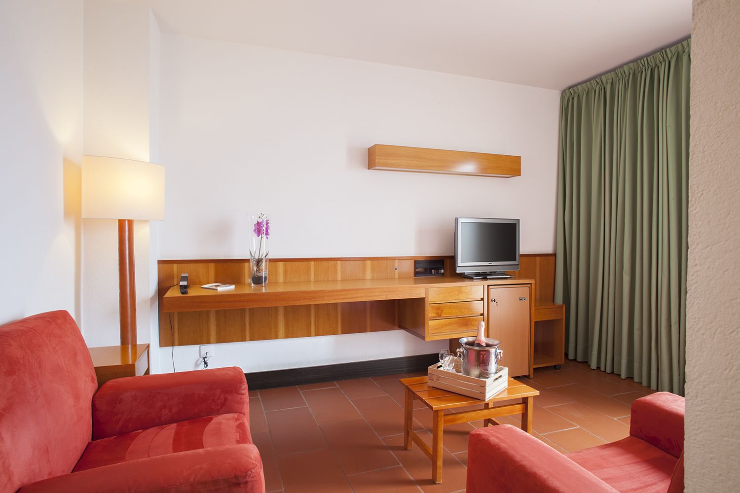 Une chambre d’hôtel avec un bureau en bois, une télévision, une petite armoire, un canapé rouge, une table basse en bois et un rideau vert.