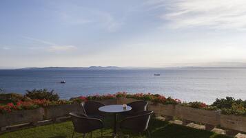 Un área de estar al aire libre con mesa y sillas, con vista a un mar tranquilo y un barco distante, además de montañas.