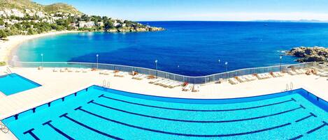 A swimming pool with multiple lanes, a beach area, and a view of the sea and distant hills.
