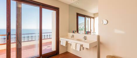 A modern bathroom with a large sliding glass door leading to a balcony, a white vanity with two sinks, and a round wooden stool.