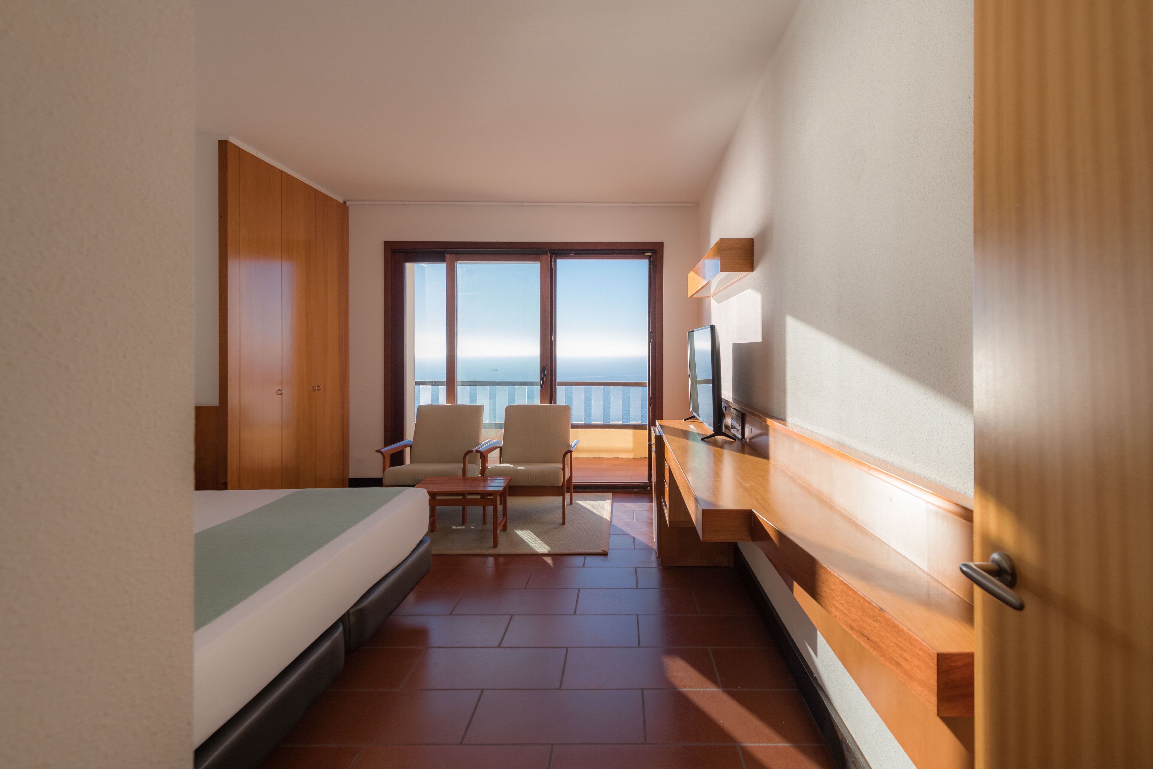 A hotel room with a bed, two chairs, a TV, and a large window with a view of the sea.