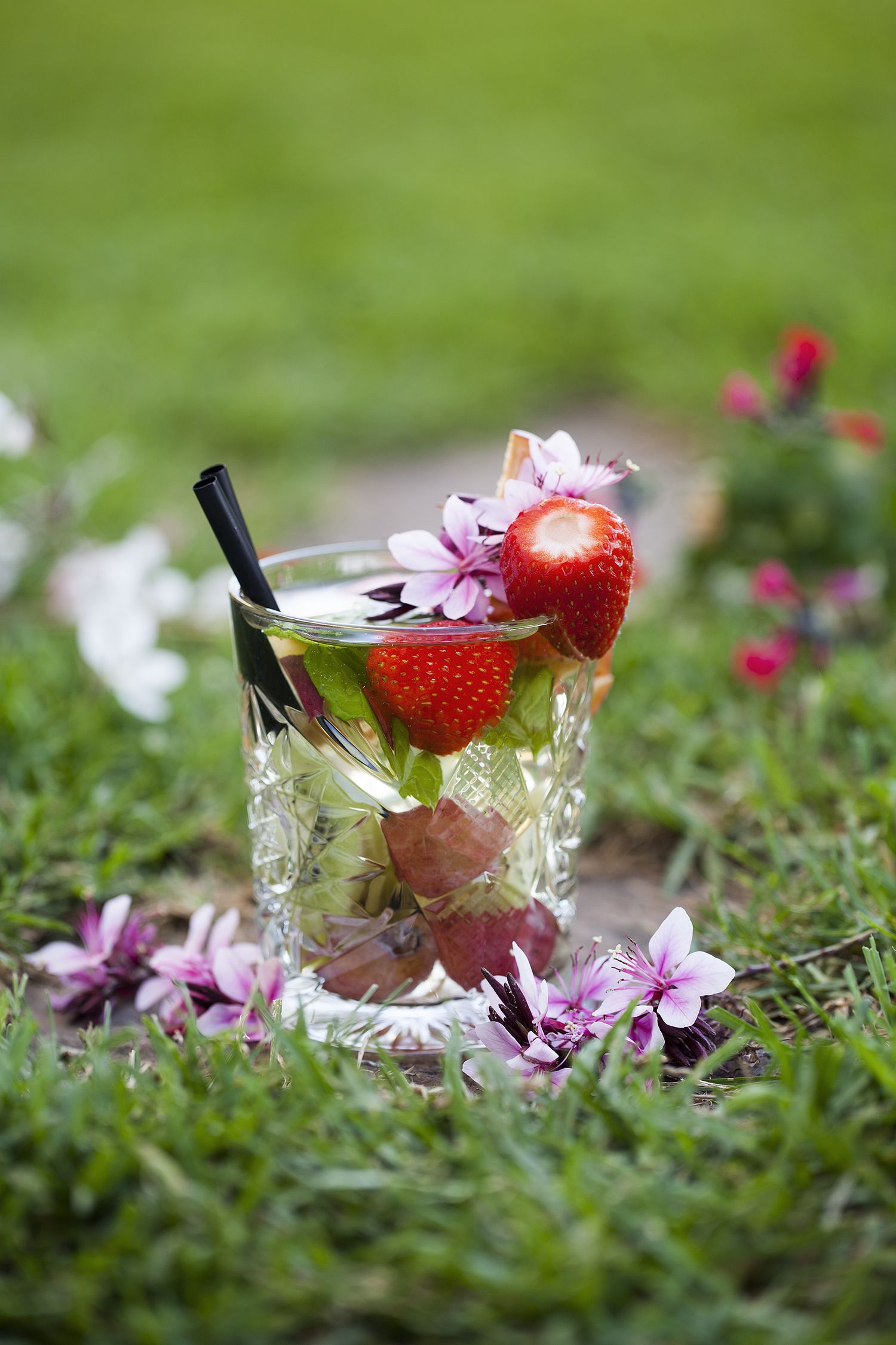 Un vaso con pajita, fresas y flores sobre un césped.