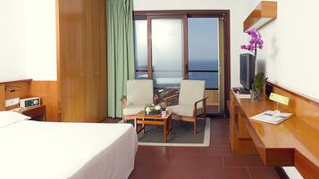 A hotel room with a bed, two chairs, a small table, a TV, and a view of the ocean.