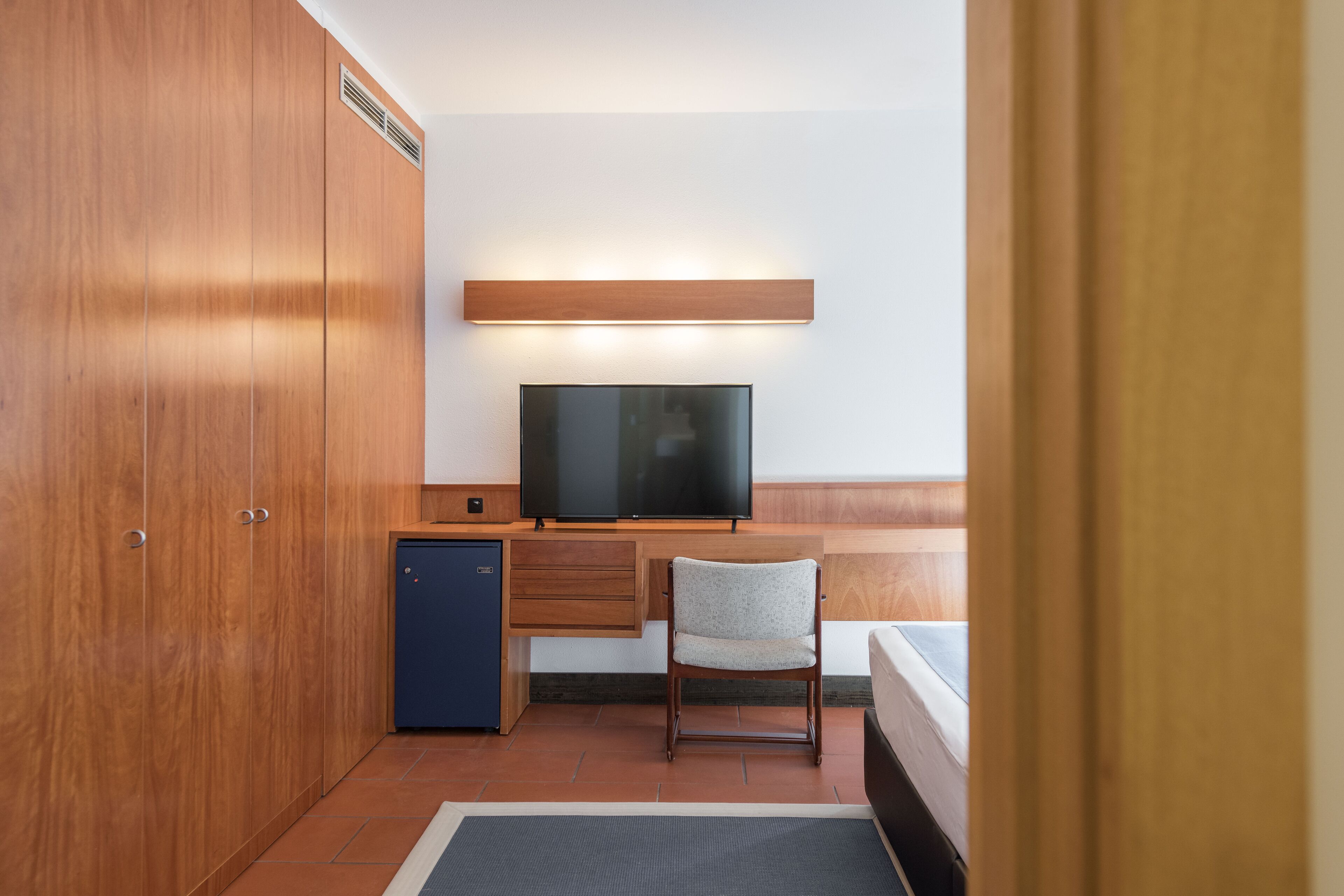 A compact hotel room with a wooden wardrobe, a small desk with a television, a chair, and a bed.