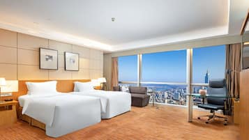 A hotel room with two beds, a desk, a chair, and large windows offering a city view.