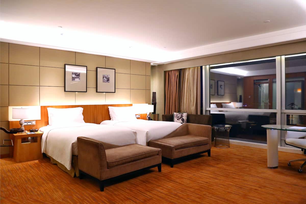 Habitación de hotel con una cama grande, dos sillones, un escritorio y vistas a la ciudad a través de un ventanal.
