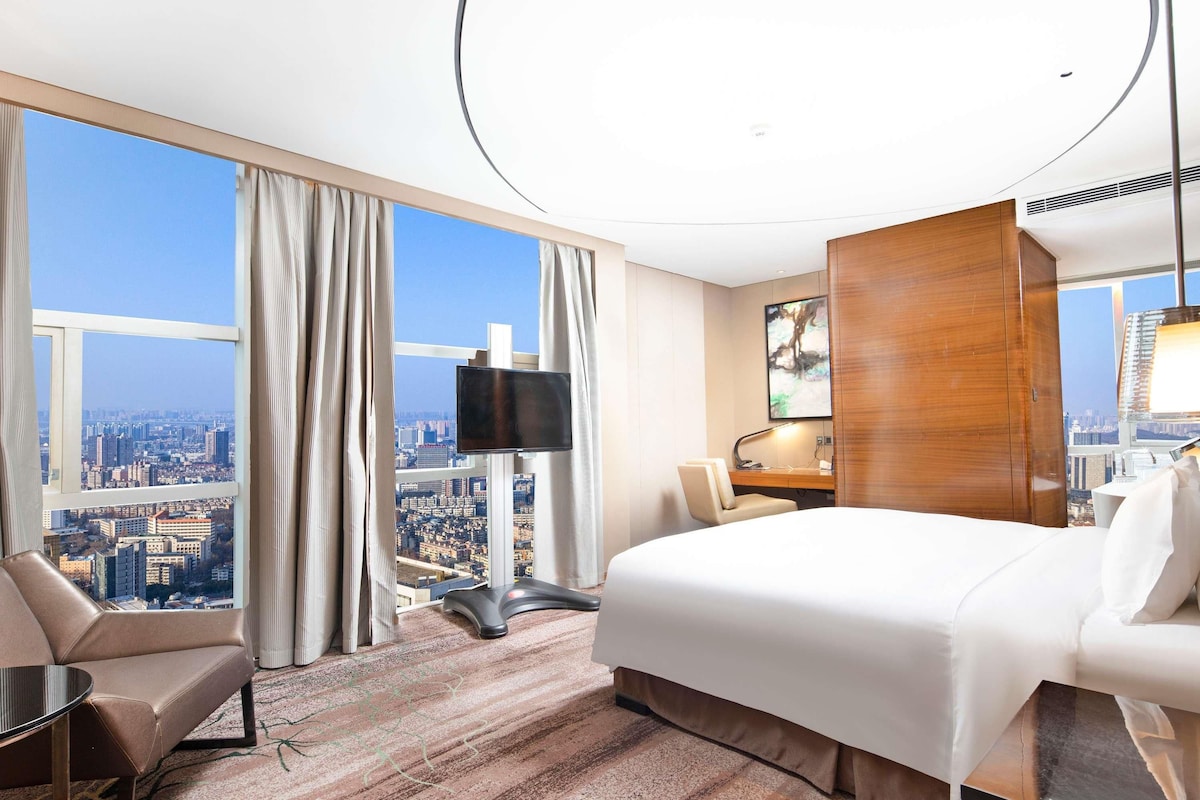 Quarto de hotel com uma cama grande, uma escrivaninha, uma cadeira e uma televisão. O quarto tem vista para a cidade através de grandes janelas.