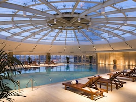 A large indoor swimming pool with a glass ceiling, wooden lounge chairs, and potted plants.