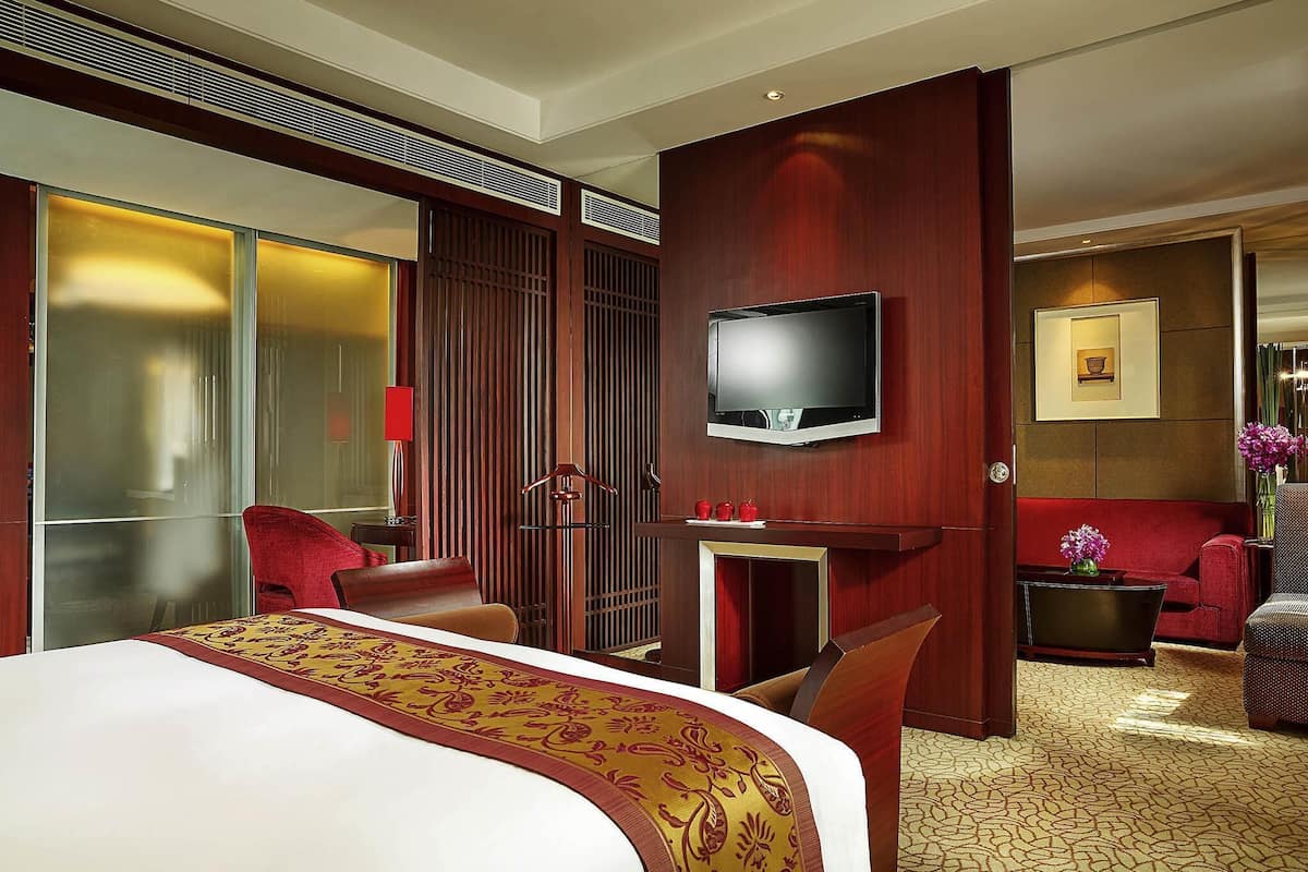 Habitación de hotel con una cama grande, un televisor montado en la pared, un sofá rojo y una mesita con un jarrón de flores.