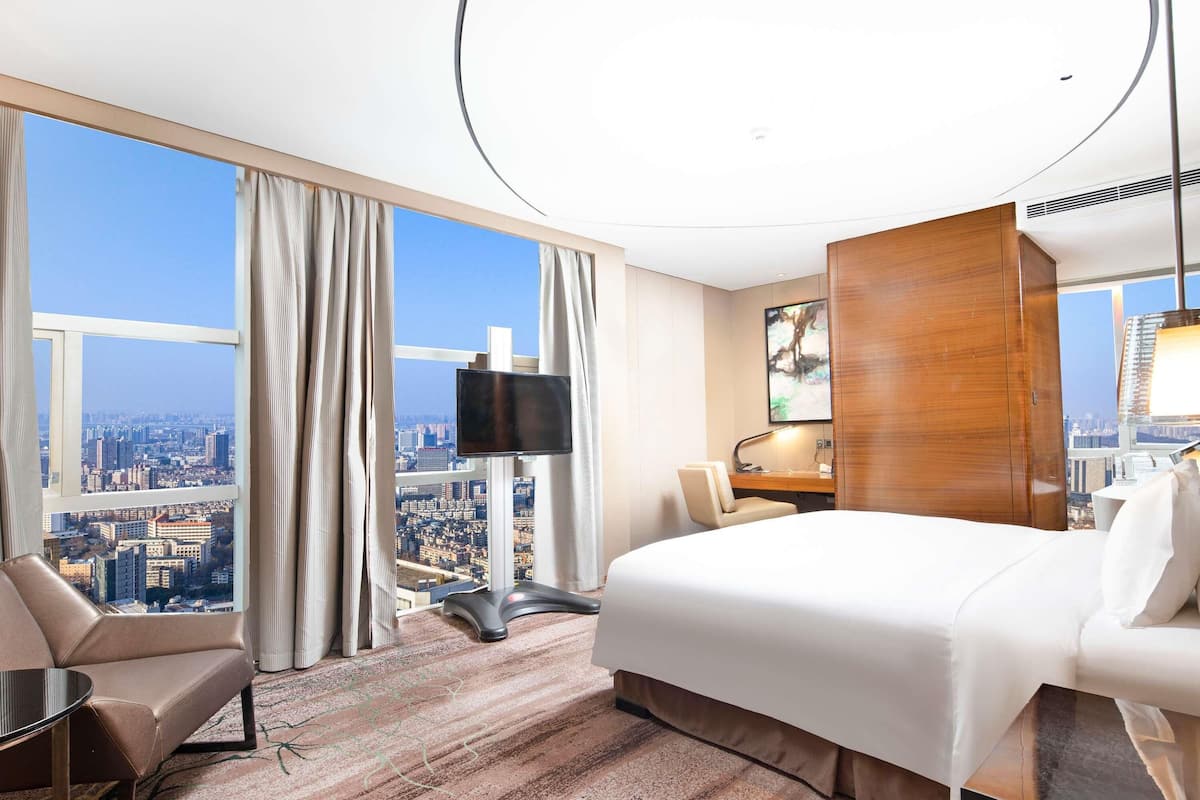 Habitación de hotel con una cama grande, un escritorio, una silla y un televisor. La habitación tiene vistas a la ciudad a través de amplias ventanas.