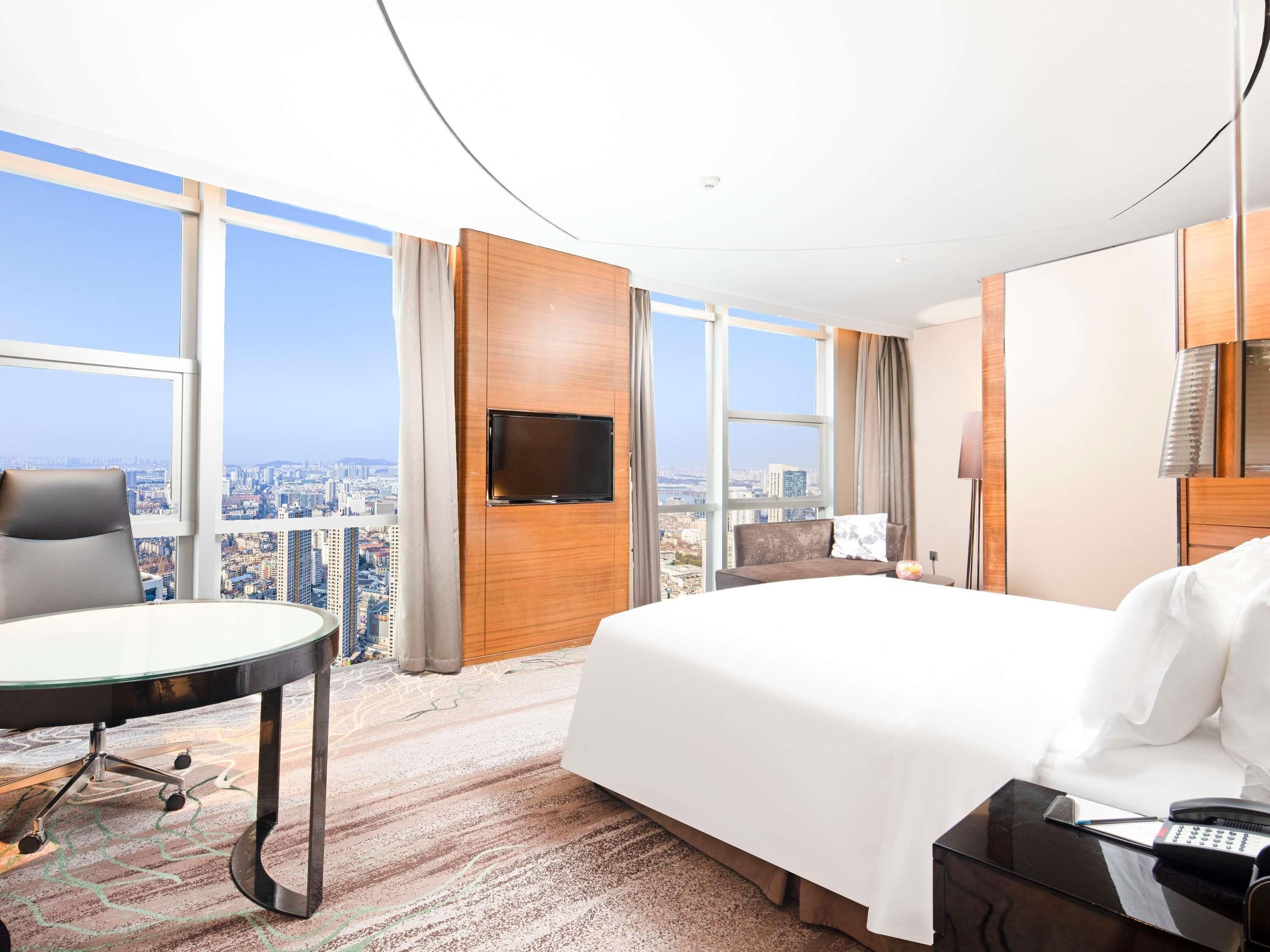 A hotel room with a large bed, a desk, and a TV. There is a view of the city through large windows.