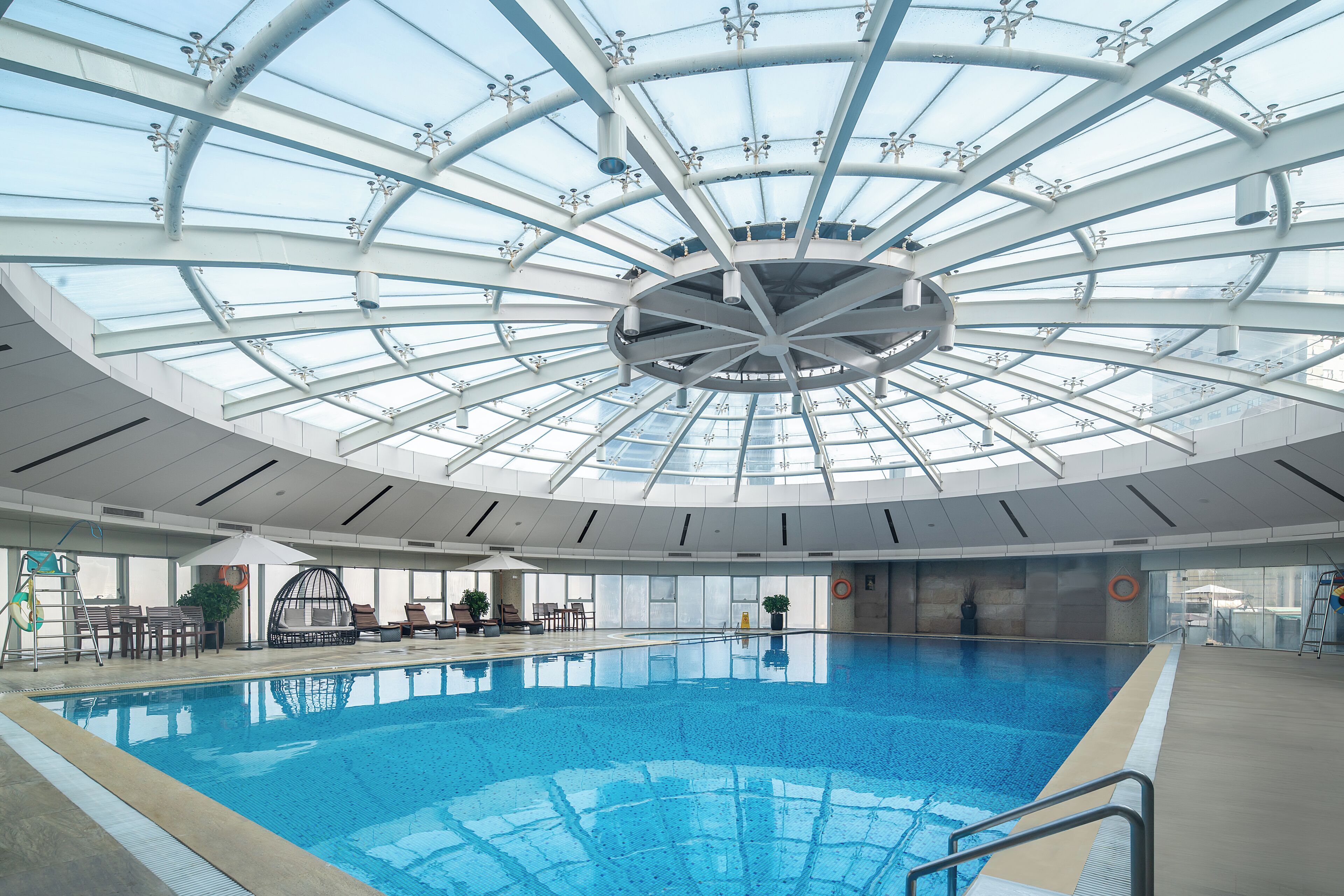 An indoor swimming pool with a glass ceiling, surrounded by lounge chairs and a sunbed.