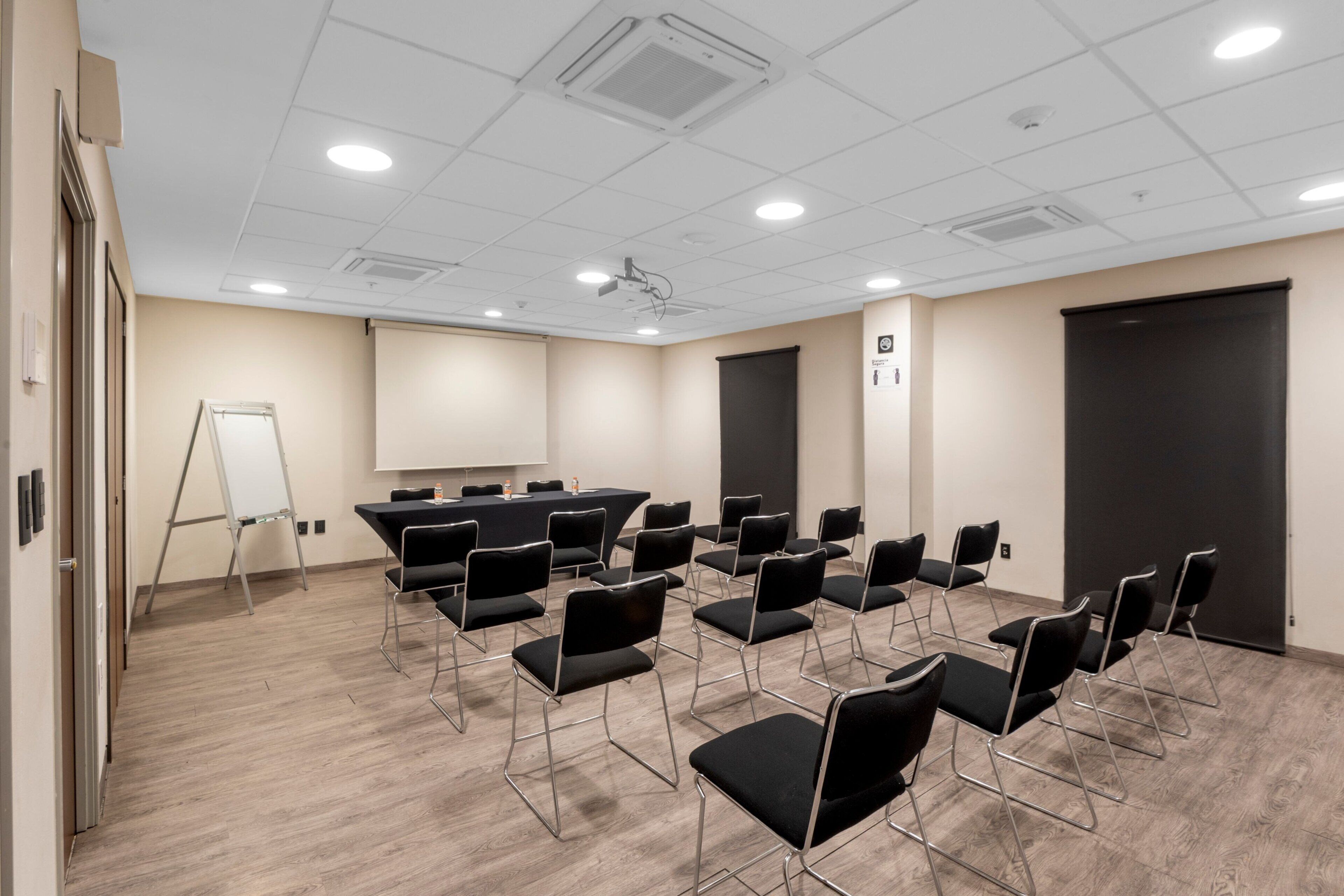 Un salón de conferencias con filas de sillas negras, una pizarra y un proyector en el techo.