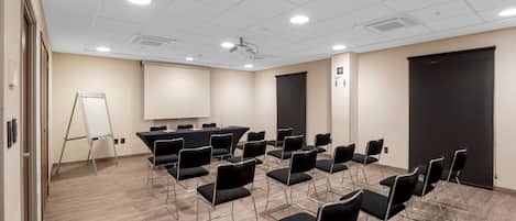 Un salón de conferencias con filas de sillas negras, una pizarra y un proyector en el techo.