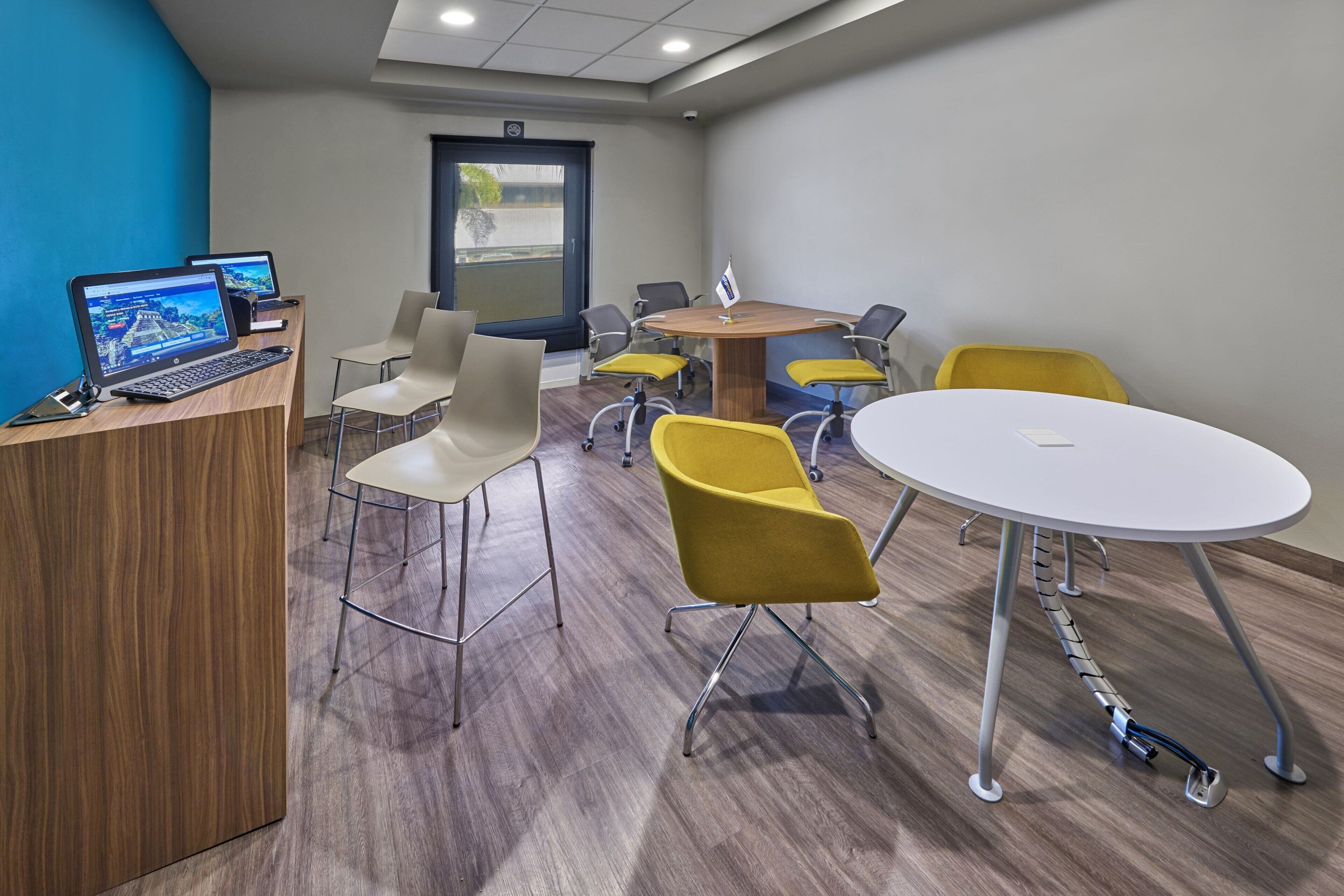 Una sala de reuniones moderna con un mostrador de recepción de madera, dos portátiles, una mesa redonda y sillas amarillas.