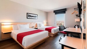 Une chambre d’hôtel avec deux lits, un bureau et une chaise. Il y a une fenêtre donnant sur un paysage urbain.