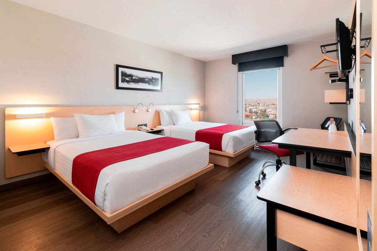 Une chambre d’hôtel avec deux lits, un bureau et une chaise. Il y a une fenêtre donnant sur un paysage urbain.