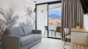 A modern living room with a grey sofa, a dining table with chairs, a small potted plant, and a view of buildings and mountains through a large window. - Hotel Merlin Resort (Adeje)