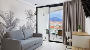 A modern living room with a grey sofa, a dining area with chairs, a small table with a plant, and a large window with a view of buildings and mountains. - Hotel Merlin Resort (Adeje)