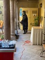Una persona con equipo de protección manejando maquinaria en un espacio exterior techado con columnas, una mesa con botellas y una planta.