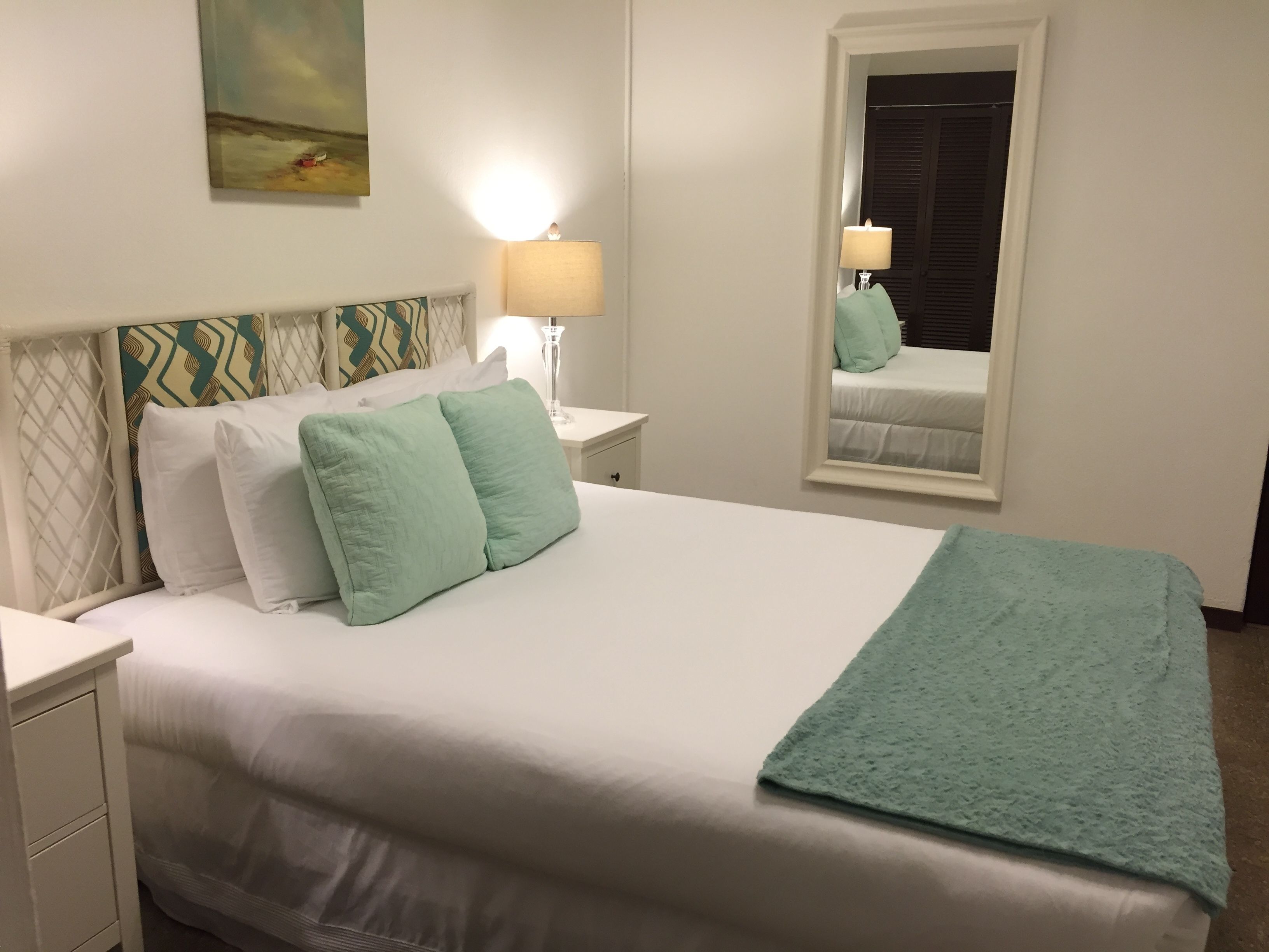 Una cama bien hecha con almohadas blancas y verde claro, una manta color turquesa y una mesita de noche con una lámpara. Un espejo refleja la cama y una ventana con persianas cerradas.