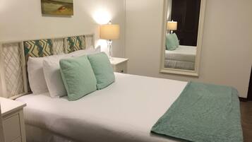 Una cama bien hecha con almohadas blancas y verde claro, una manta color turquesa y una mesita de noche con una lámpara. Un espejo refleja la cama y una ventana con persianas cerradas.